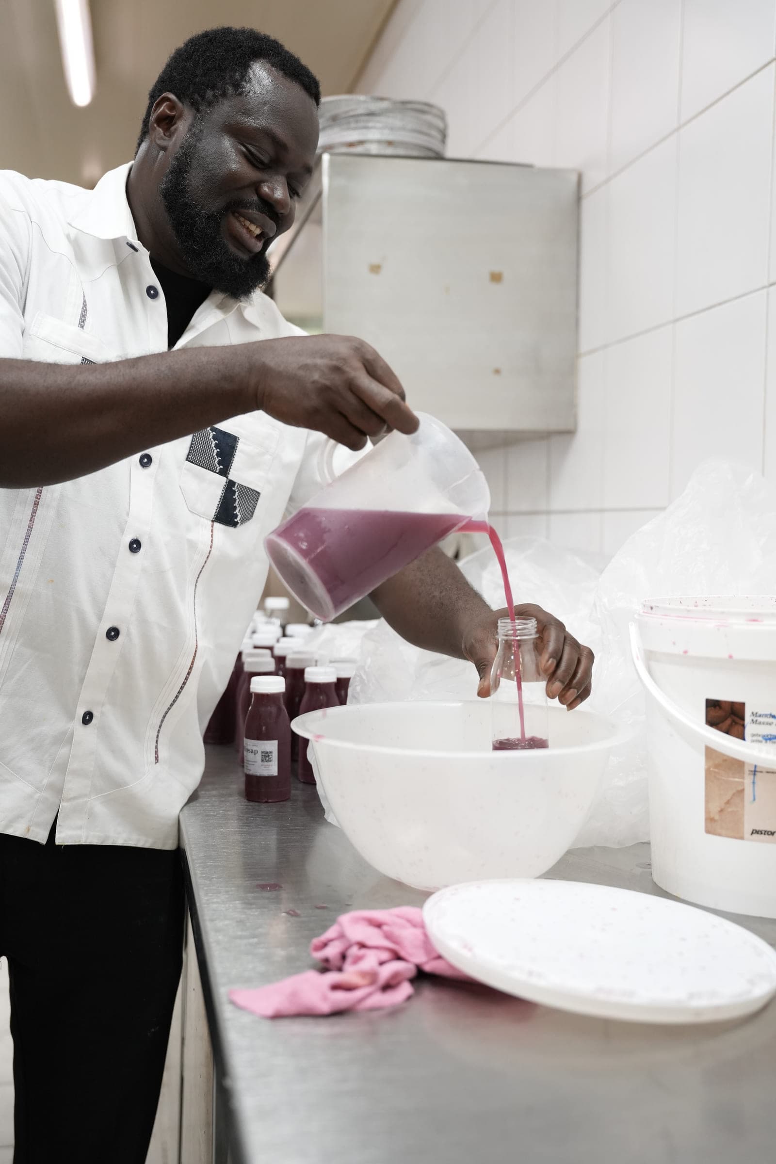 A man in a white kitchen jacket pours bissap from a measuring jug into a bottle held over a mixing bowl; filled and labelled bottles wait beside him.