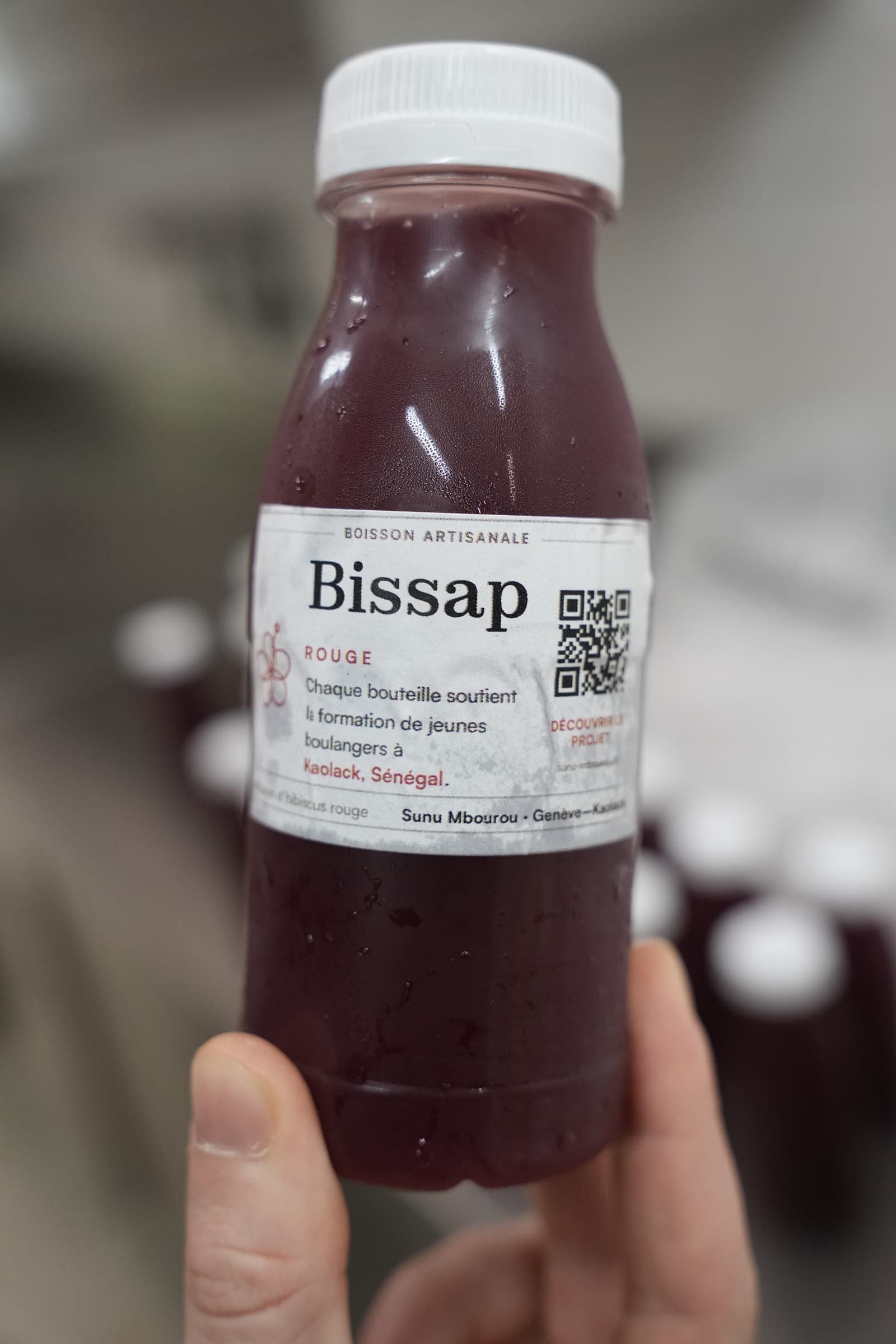 A hand holds a bottle of red bissap. The label carries a QR code and, in French, the sentence “Every bottle supports the training of young bakers in Kaolack, Senegal”.
