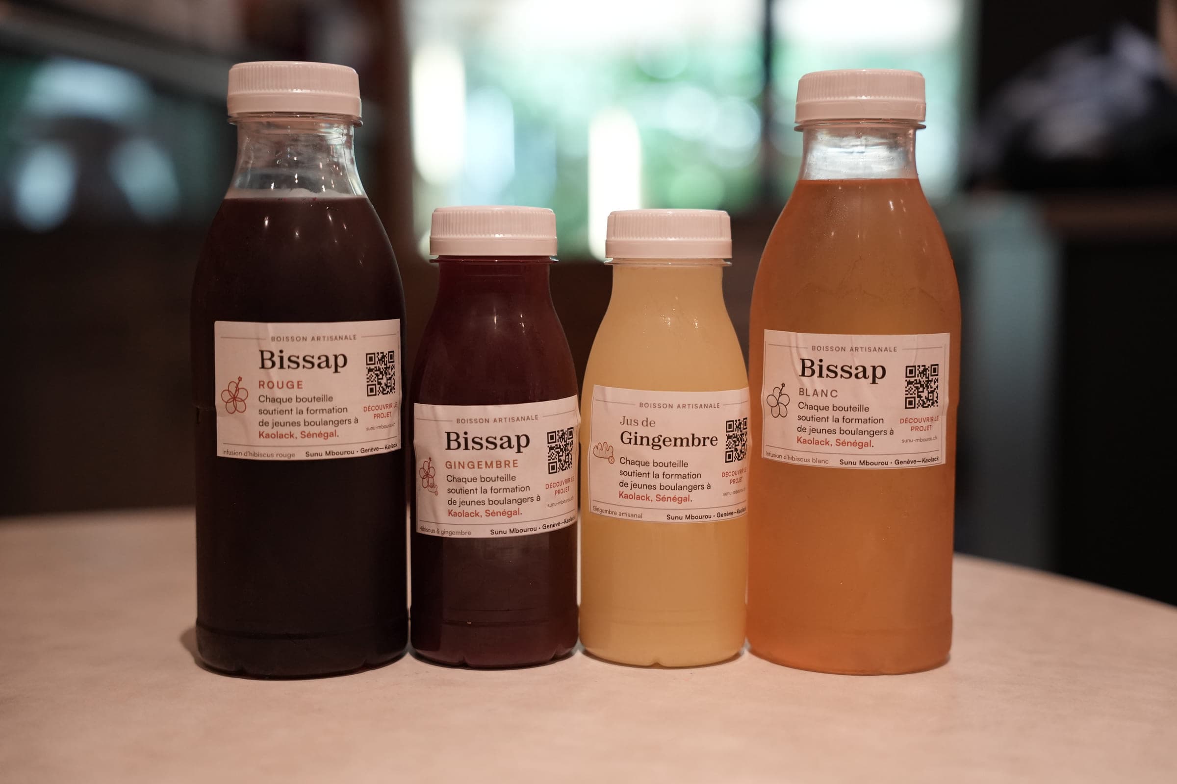 Four bottles lined up on a table: red bissap, ginger bissap, ginger juice and white bissap, each labelled and carrying a QR code.