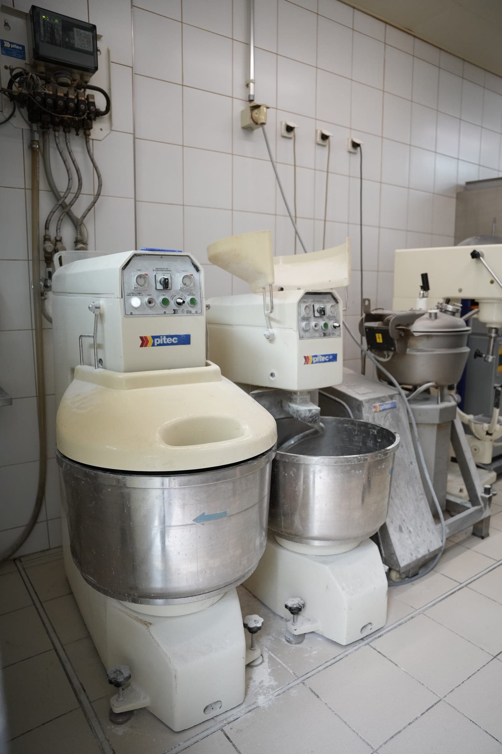 Two spiral mixers lined up against a tiled bakehouse wall, their stainless-steel bowls visible, a third machine behind.