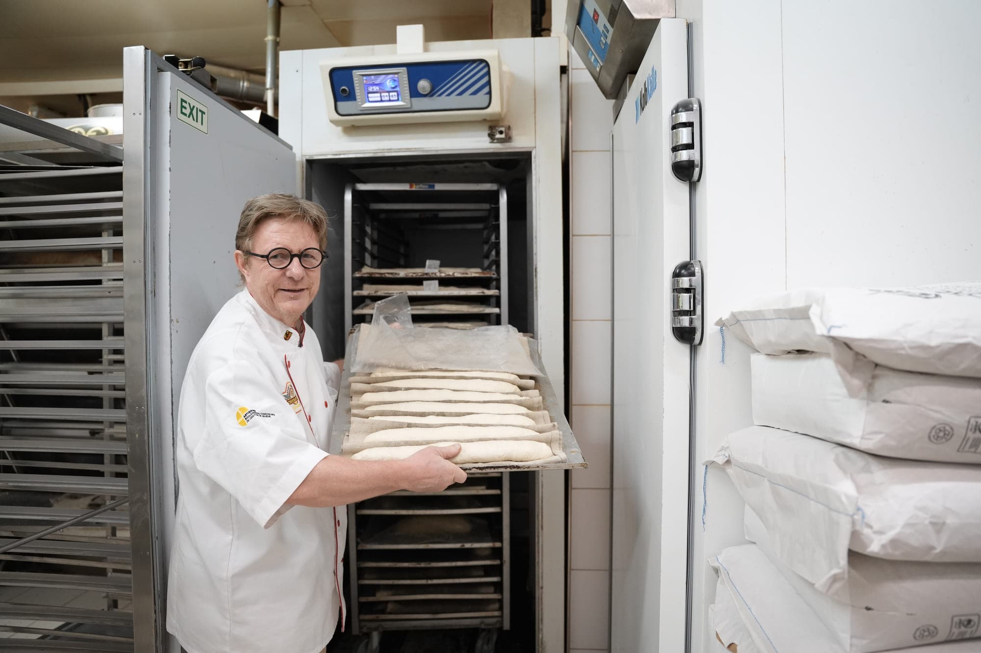 A baker slides a board of raw baguettes, still covered with a cloth, into a rack.