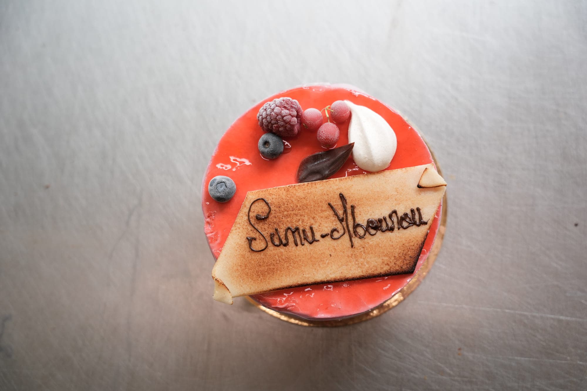 A round entremets glazed in red, topped with red fruit and a meringue, carrying a marzipan plaque on which the project’s name is written in chocolate.