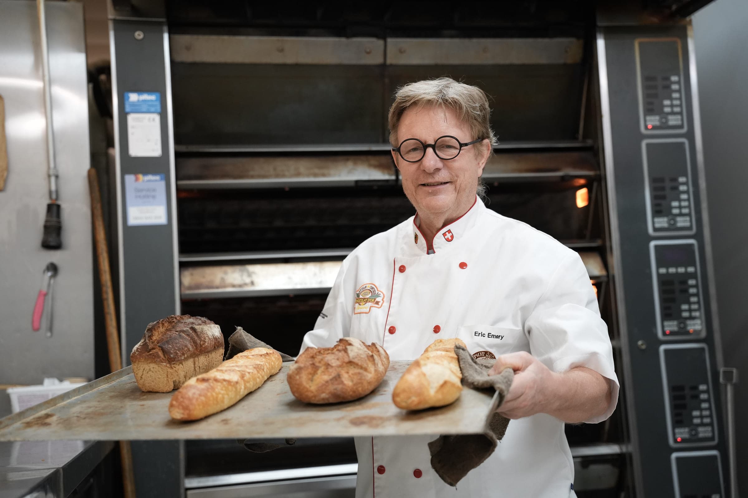 A baker in a white jacket holds, with both hands, a wooden board carrying four baked loaves and baguettes, in front of a deck oven with its windows lit.