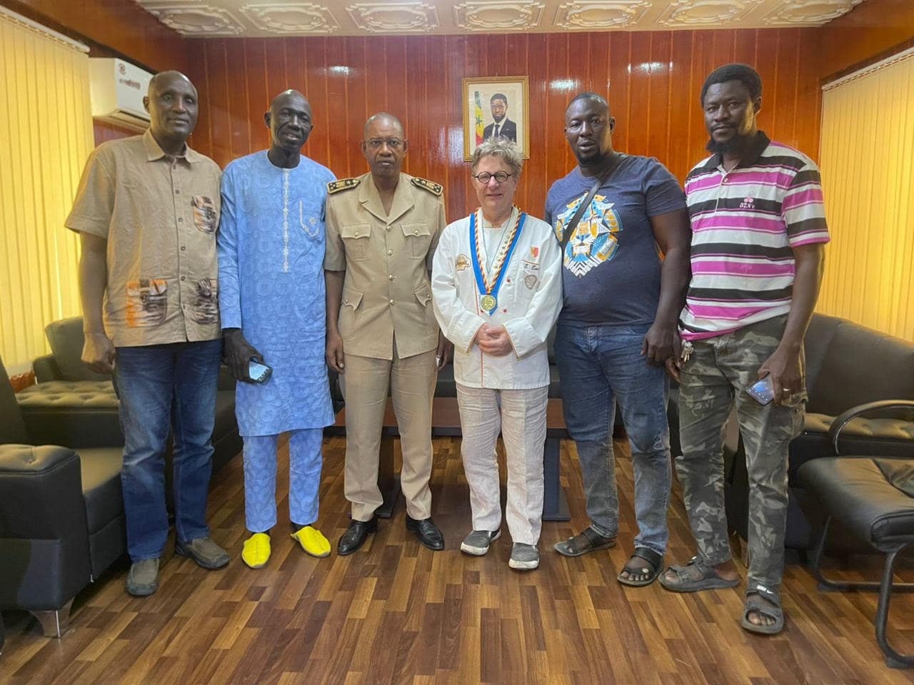 Six personnes posent dans un bureau officiel au Sénégal. Au centre, Eric Emery porte une veste blanche de boulanger et une médaille professionnelle ; à sa droite, un représentant de l’administration en uniforme.